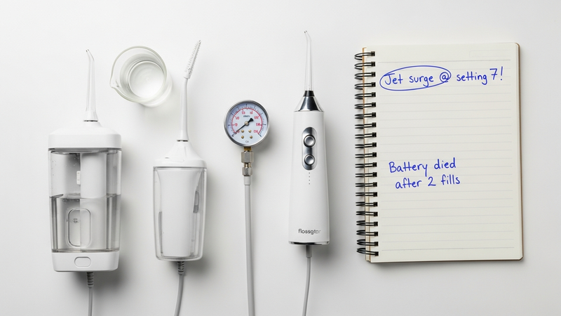 An overhead shot of a practical testing station: two flossers, beakers of water, a pressure gauge, and a notebook with candid observations like "Jet surge @ setting 7!" and "Battery died after 2 fills."