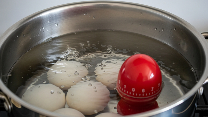 A first-person perspective shot looking down into a stainless steel pot. Several eggs and a vibrant red (starting color) egg timer are submerged in clear, bubbling water. Steam rises gently.