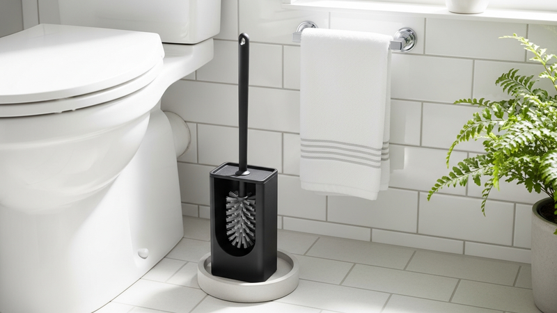 A lifestyle shot in a bright, clean bathroom. A stylish silicone brush system sits neatly beside the toilet. The scene looks intentional and sanitary, not like an embarrassing necessity stuffed in the corner.